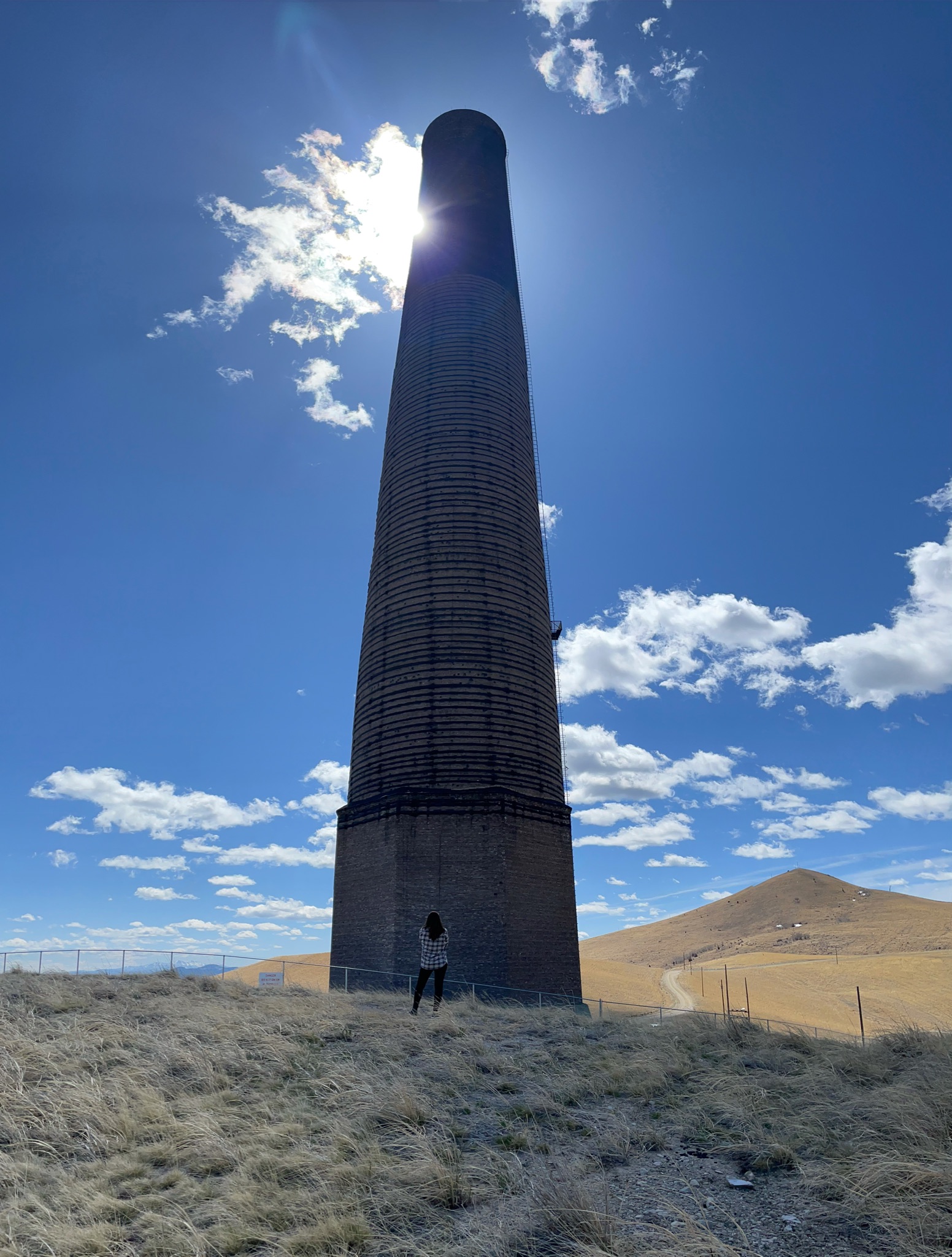 The Anaconda Smelter, in Anaconda, Montana is pictured with Melissa standing at its base. The smelter stands tall in front of a blue sky.